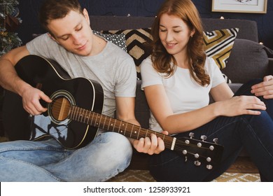 Young Couple Sitting On The Floor At Home Near The Christmas Tree With Gifts And The Guy Playing The Guitar, New Year's Eve