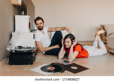 Young Couple Sitting On The Floor, Listening To Music On A Vinyl Player With Different Vinyl Records.