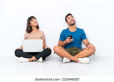 Young Couple Sitting On The Floor Holding Pc And Mobile Phone Isolated On White Background Looking Up While Smiling