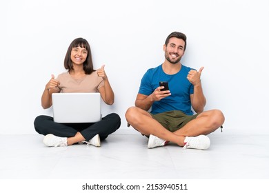 Young Couple Sitting On The Floor Holding Pc And Mobile Phone Isolated On White Background Giving A Thumbs Up Gesture With Both Hands And Smiling