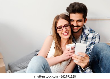 Young Couple Sitting On The Floor Of Empty Apartment Looking At Smart Phone. Move In To New Home.