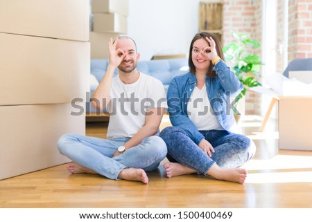 Young couple sitting on the floor arround cardboard boxes moving to a new house doing ok gesture with hand smiling, eye looking through fingers with happy face.