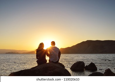 Young couple sitting on a rock near the sea and watching the sun