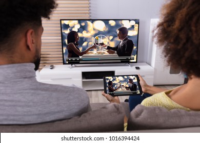 Young Couple Sitting On Sofa Connecting Television Channel Through WiFi On Digital Tablet