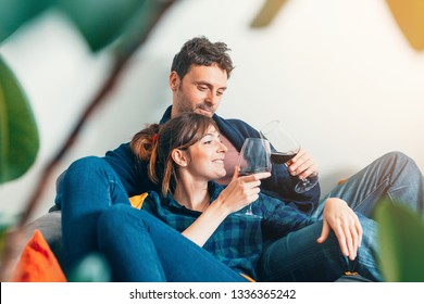 Young Couple Sitting On Sofa At Home, Toasting And Smiling - Home Life, Living Together, And Domestic Partnership Concept