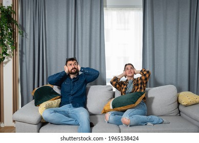 Young couple is sitting on a sofa in their apartment looking up and holding their hands to plug their ears as a neighbor upstairs is having a party and playing loud music or renovating the apartment 