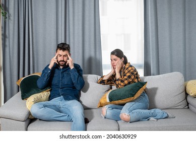 Young Couple Is Sitting On A Sofa In Their Apartment And Holding Their Hands To Plug Their Ears As A Neighbor Upstairs Is Having A Party And Playing Loud Music Or Renovating The Apartment 