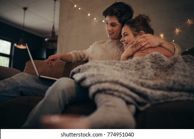 Young Couple Sitting On Sofa At Home In Winter And Using Laptop Computer. Cozy Couple In Hygge House With Laptop.