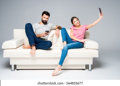 Young Couple Sitting On White Couch And Using Smartphones Isolated On Grey