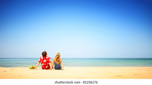 Young couple sitting on yellow sand by blue sea and looking to a clear sky