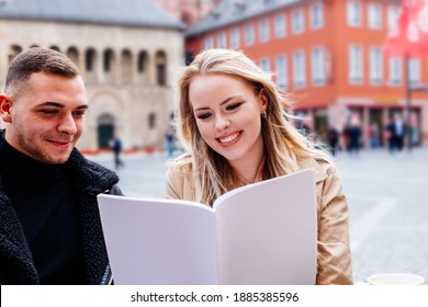 Young Couple Sitting Outside In Cafe Happily Reading Magazine With Blank Cover As Mock Up.