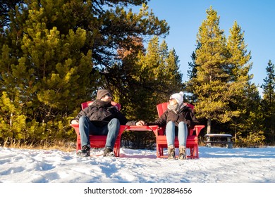 Young Couple Sitting In Red Adirondack Chairs And Looking At Each Other, At Maligne Lake, Canada