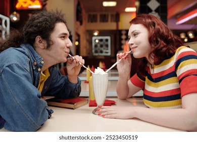 Young couple sitting in retro diner sharing milkshake while smiling and looking at each other without a care in the world enjoying the nostalgic setting, adding to the romantic atmosphere - Powered by Shutterstock