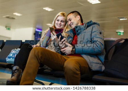 Young couple sitting in the terminal waiting room and waiting for departure, she is texting and connecting with her smartphone