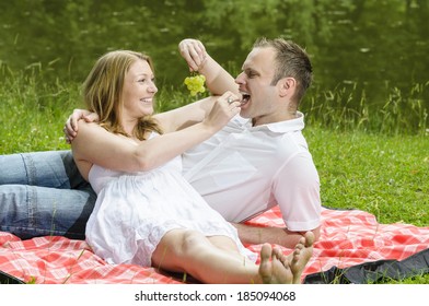 Young couple sitting together in a summer park and have a picnic on a checkered picnic blanket. They feed each other with treats.