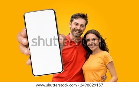 A young couple smiles at the camera while holding a smartphone with a blank screen in front of a bright yellow background.