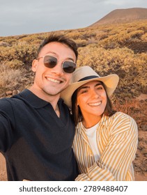Young couple smiling at the camera, capturing a travel selfie together while exploring a dry volcanic landscape at sunset, enjoying their summer vacation