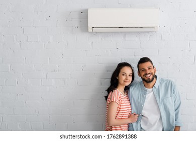 Young couple smiling at camera while standing near air conditioner on wall at home