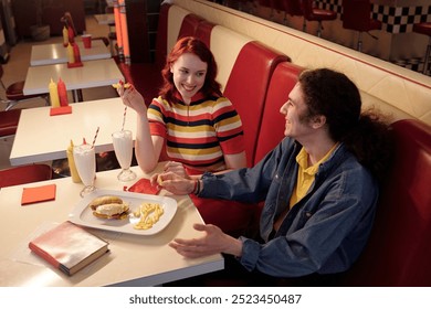 Young couple smiling and enjoying their meal in a retro diner booth, with milkshakes, fries, and a burger on table, having a pleasant conversation - Powered by Shutterstock