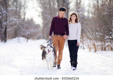 Young Couple Smiling And Having Fun In Winter Park With Their Husky Dog.