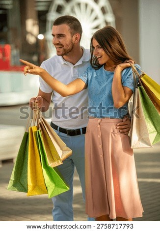 Young couple smiling and pointing while holding colorful shopping bags outside a mall.