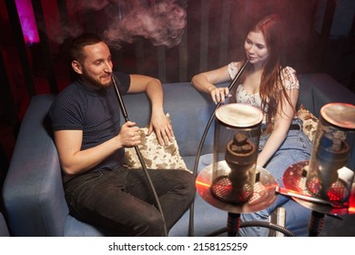 a young couple are smiling and sitting in a hookah lounge on the couch and smoking a hookah, releasing smoke