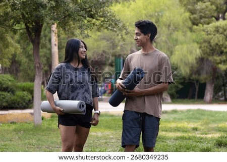 Young couple smiling and talking while carrying rolled yoga mats in a green park, preparing for an outdoor exercise session and promoting a healthy lifestyle