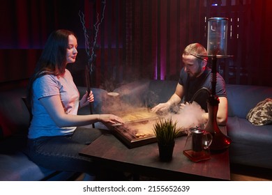 a young couple smokes a hookah in an atmospheric hookah lounge in a relaxed atmosphere, wooden backgammon is in front of him, they play