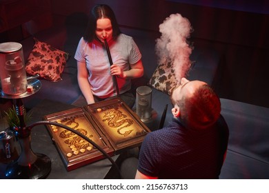 young couple smoking hookah in atmospheric hookah bar. sit freely on sofas, play backgammon and blow smoke