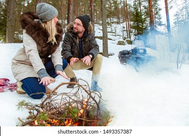 Young Couple And Snowmobile In Winter Forest.Lovers Make A Fire. Friends And A Snowmobile At The Winter Bonfire.People Around The Campfire Are Smiling. Outdoor Activities During The Christmas Holidays