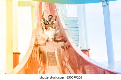 Young Couple Splashing Into A Waterslide At Aqua Park For Summer Vacation - Multiracial People Having Fun Outdoor - Travel And Love Concept - Focus On Them - Radial Blue And Yellow Filters Editing 