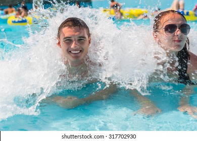 Young Couple Splashing In Shallow Waves Of Wave Pool On Sunny Day - Young Couple Cooling Off In Waves In Busy Pool At Resort Water Park