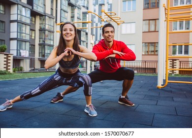 Young Couple In Sports Outfit Doing Morning Workout Outdoors. Young Mixed Race Man And Woman Stretching They Muscle Before Running On Street Sports Ground. Staying Fit And Healthy Concept