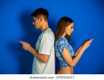 Young couple standing back to back while using smartphones on blue background.