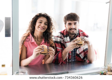 A young couple is standing by a mobile snack bar in the street. They ordered two juicy, fragrant burgers and eat them with appetite. They are smiling