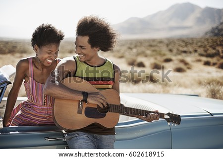 A young couple standing by a pale blue convertible on the open road, the man playing a guitar