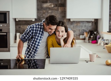 A young couple standing in a kitchen at home, using laptop.
