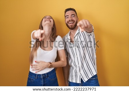 Young couple standing over yellow background laughing at you, pointing finger to the camera with hand over body, shame expression 