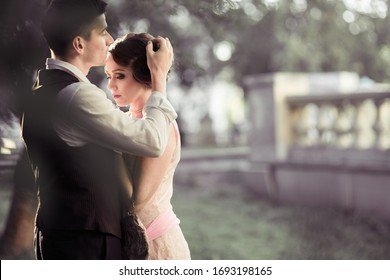 A Young Couple Stands Against A Backdrop Of Large Trees And A Fence. A Man Adjusts The Hair On A Woman's Head. Historical Reconstruction