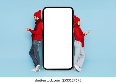 A young couple stands back to back wearing red sweaters and hats, focused on their smartphones. They are positioned next to a large blank screen, suggesting a playful or social setting. - Powered by Shutterstock