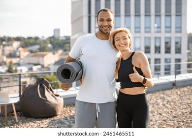 Young, couple stands joyfully on rooftop, exuding happiness and health after fitness session. Man holds rolled-up yoga mat, while woman gives thumbs-up, both dressed in sportswear on urban backdrop. - Powered by Shutterstock