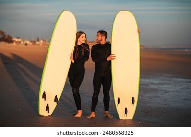 A young couple stands on a sandy beach at sunset, holding their surfboards. The sun is setting in the background, and the sky is a beautiful orange and pink. - Powered by Shutterstock