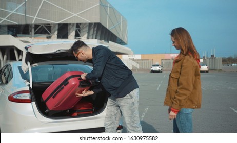 Young Couple With Suitcases Approaching Car. Man In Casual Clothes Putting Luggage In Trunk. White Car. Travelers Near Airport.