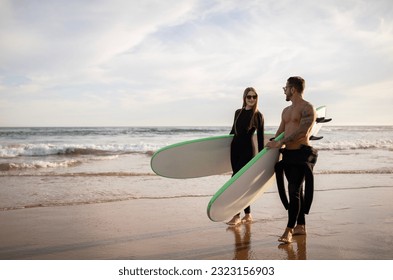 Young couple of surfers walk with surfboards on the beach at sunset, happy lovers wearing wetsuits going to surf together, man and woman enjoying water sports and active lifestyle, copy space - Powered by Shutterstock