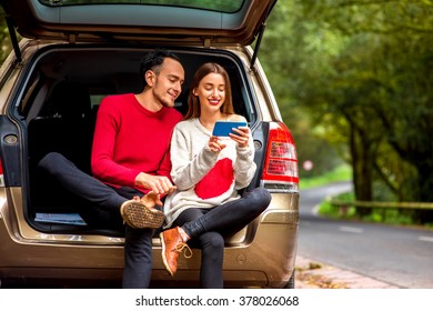 Young Couple In Sweaters Sitting Together In The Car Trunk With Smart Phone On The Roadside In The Forest. Young Family Traveling By Car