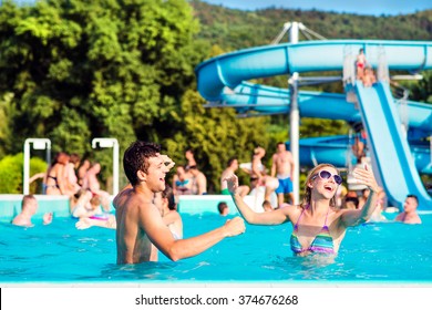 Young Couple In Swimming Pool On Sunny Day. Water Slide.