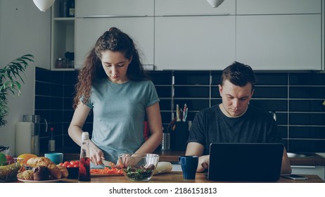 young couple at table, man is sitting working on laptop, he is concentrated and pensive, beautiful curly woman is standing cooking cutting vegetables for salad, she is seriuos