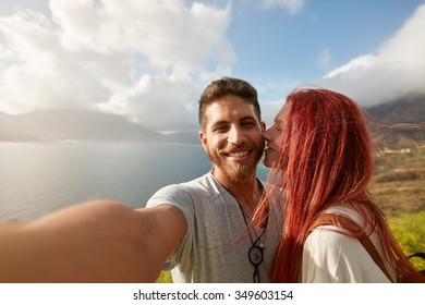 Young Couple Taking A Selfie Outdoors. POV Shot Man Holding A Camera And Taking A Self Portrait With Woman Kissing Him.