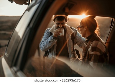 Young couple talking, drinking a coffee watching the sunset from their car. Couple road travel together - Powered by Shutterstock