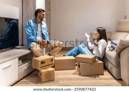 Young couple talking and smiling while unpacking cardboard boxes in new home, planning the arrangement of furniture